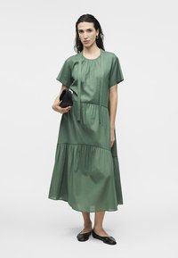 Γυναίκα με χαλαρό, πράσινο, στρωματοποιημένο μίντι φόρεμα, κρατώντας ένα μαύρο τσαντάκι χειρός και φορώντας μαύρα φλατ παπούτσια, στέκεται μπροστά σε λευκό φόντο.