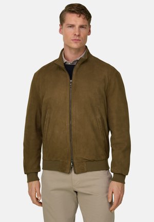 Uomo che indossa una giacca in camoscio marrone con zip sopra una camicia beige e pantaloni chiari, in piedi contro uno sfondo semplice.