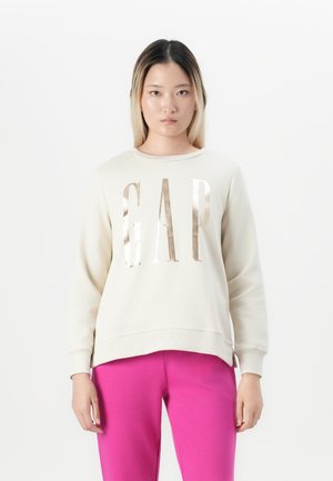 Young woman with long dark and blonde hair wearing a cream GAP sweatshirt and bright pink pants, standing against a white background.