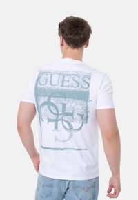 Camiseta de algodón blanca con un gran estampado gráfico en teal con la palabra "GUESS" y formas abstractas en la parte posterior. Diseño de corte clásico.