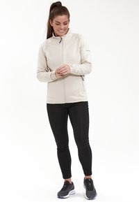 Beige fleecejacka med hög krage och frontdragkedja, kombinerad med svarta figurnära byxor och mörka sportskor. Slät textur, minimalistisk design.