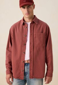 Rode katoenen shirt met knopenfront, kraag en enkele zak. Draag het over een lichtroze shirt, gecombineerd met lichtblauwe spijkerbroek.