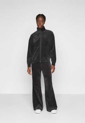 Zwarte corduroy trainingspak met een ritsjack en wijde broekspijpen. Voorzien van ribbelstructuur en zijzakken, gecombineerd met witte sneakers.