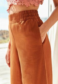 Pantalons bruns, légers, à jambes larges avec une taille élastique et des poches latérales, fabriqués dans un tissu doux au toucher lisse.