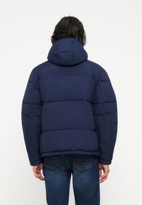 Chaqueta acolchada azul marino con capucha, con secciones acolchadas y una textura exterior suave. Combinada con jeans de mezclilla azules ajustados.