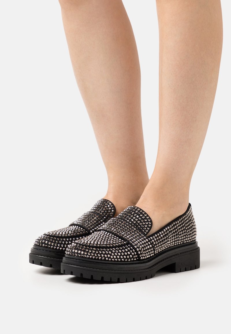 Mocassins noirs à semelles épaisses recouverts de clous argentés portés pieds nus sur un fond blanc uni.