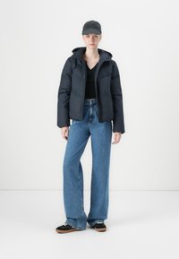 Black padded jacket with a hood, paired with a black top, flared blue jeans, and black sneakers. Accessories include a dark cap.