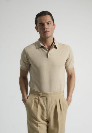 Camiseta polo de manga corta beige con cuello plano y tapeta de tres botones, combinada con pantalones de tela beige claros con pliegues y bolsillos laterales.