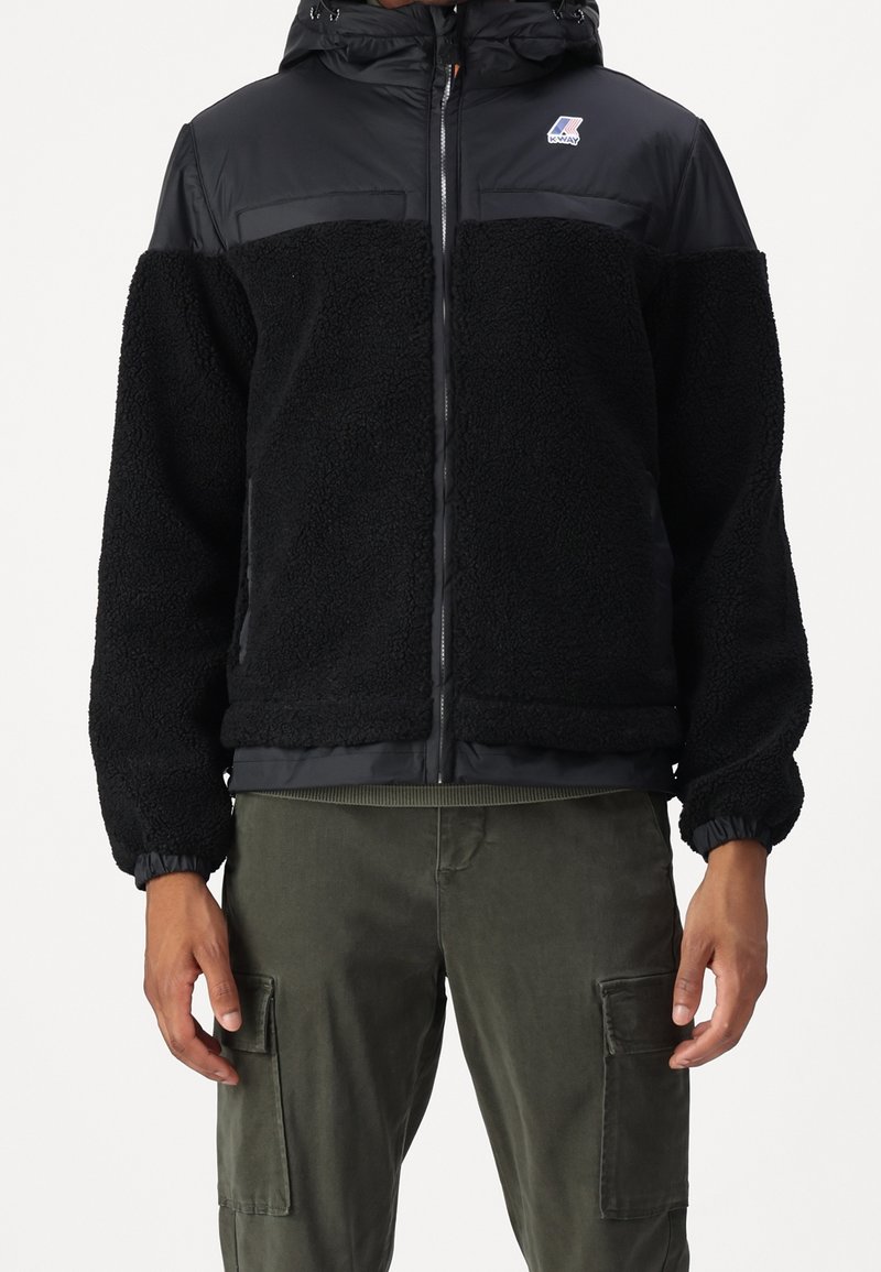 Homme portant une veste zippée noire texturée avec capuche et un pantalon cargo vert foncé sur fond blanc.