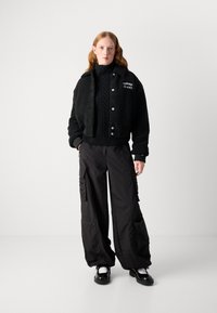 Veste noire courte avec un extérieur texturé, associée à un pantalon cargo noir ample avec des poches zippées, portée avec des chaussures noires.