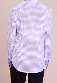 Person som bär en åtsittande långärmad lavendel- och vitrutigt skjorta, sedd bakifrån, i kombination med svarta byxor.