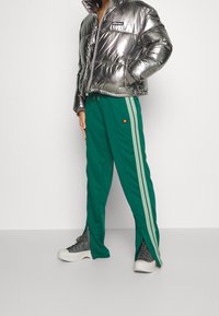 Metalická stříbrná prošívaná bunda na zip s logem, doplněná zelenými tepláky s vertikálními světle zelenými pruhy a detaily na zip.