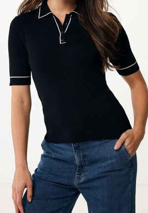 Kobieta ubrana w czarną prążkowaną koszulkę polo z krótkim rękawem i białą lamówką oraz niebieskie dżinsy, z jedną ręką w kieszeni, stoi na białym tle.