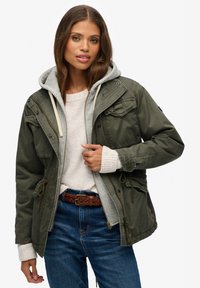 Olivenrgrøn jakke med lynlåslukning foran, lommer og snørelukning i taljen, lagdelt over en grå hættetrøje og cremefarvet sweater. Blå jeans, flettet brunt bælte.