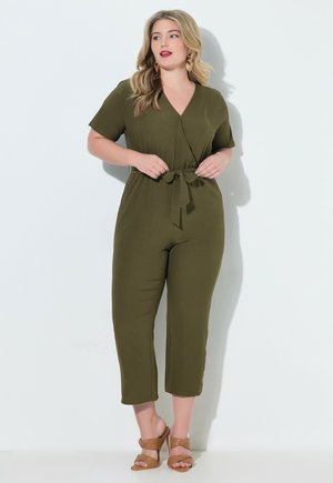 Nainen pukeutuneena oliivinvihreään lyhythihaiseen haalariin, jossa on vyötärösolki, ja hänellä on ruskeat korkokengät, seisoo valkoista taustaa vasten.