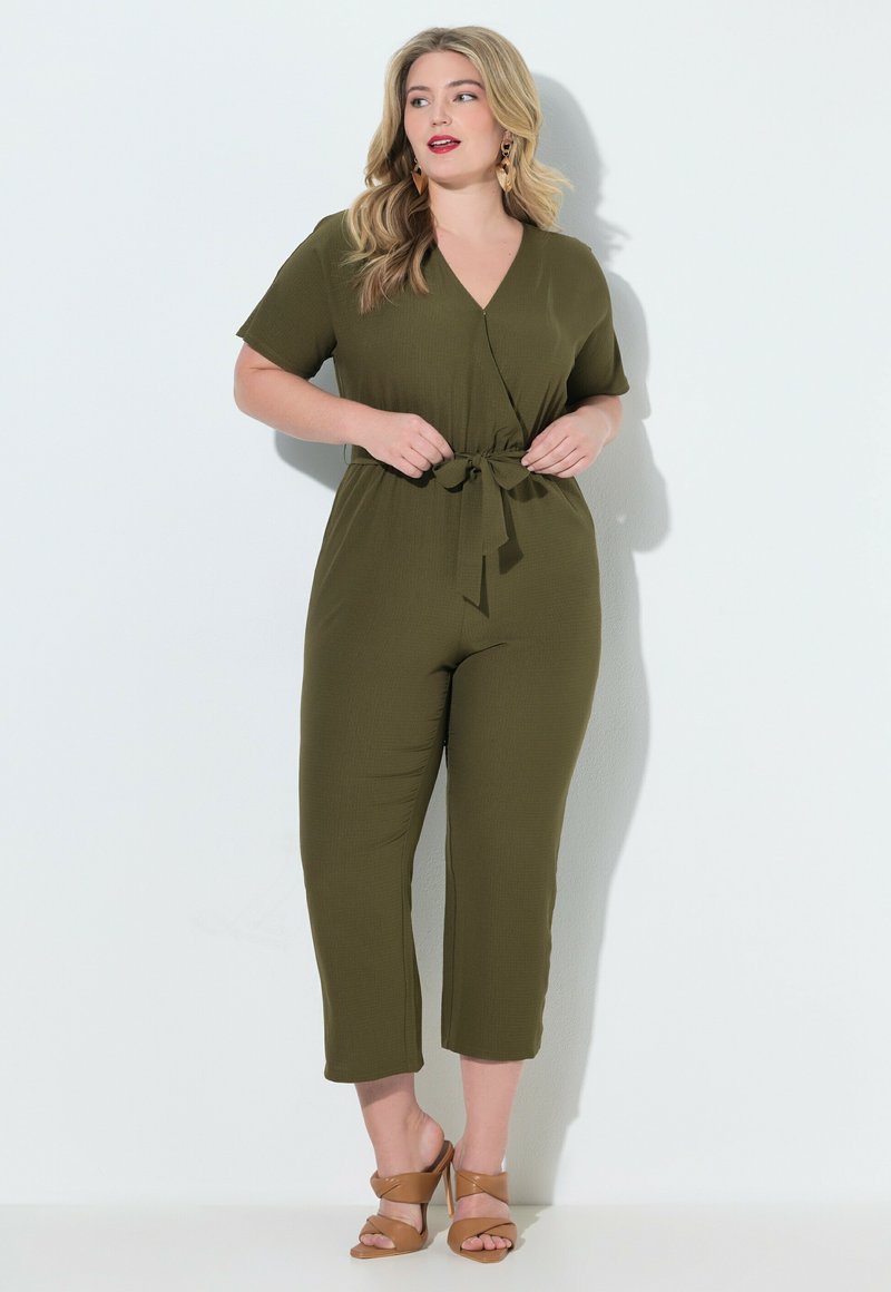 Mujer vestida con mono de manga corta color verde oliva con cinturón en la cintura y sandalias de tacón alto color beige, de pie frente a un fondo blanco.