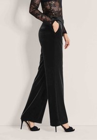 Vrouw in zwarte wijde fluwelen broeken en hoge open teen schoenen, met hand in de zak en een lange mouwen top van kant.