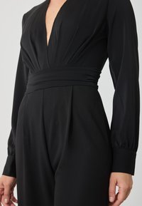 Combinaison noire avec manches longues, décolleté en V profond, devant plissé et large taille, fabriquée en tissu lisse et texturé.
