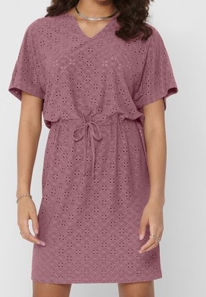 Kobieta w sukience z ażurowej koronki w kolorze mauve, z krótkimi rękawami, dekoltem w serek oraz wiązaniem w talii, dopełniona pierścionkami i bransoletkami.