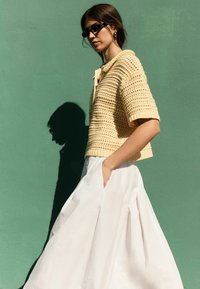 Mujer con gafas de sol negras, top corto amarillo de punto y falda blanca, de pie contra una pared verde con la mano en un bolsillo.