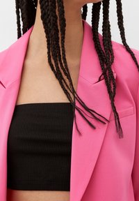 Veste rose vif à revers crantés, associée à un crop top noir côtelé. Les cheveux sont longs et tressés.