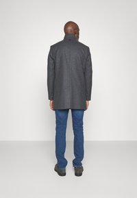 Cappotto grigio in misto lana con collo alto, taglio dritto e due tasche laterali, abbinato a jeans in denim blu e scarpe grigie.