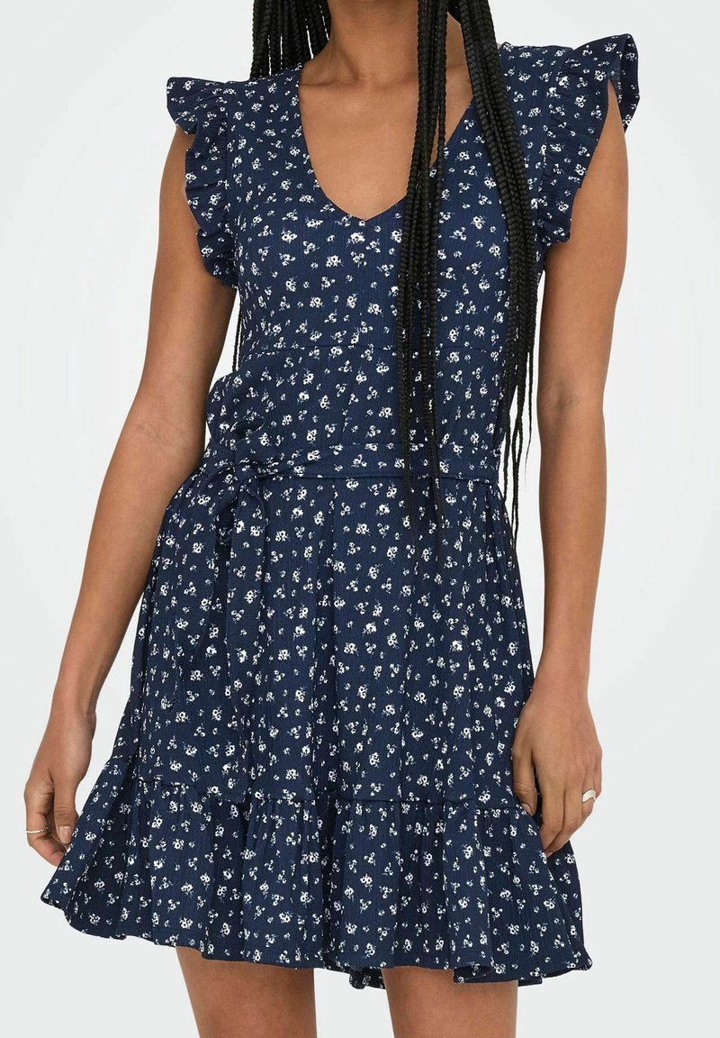 Femme portant une robe sans manches bleu marine avec imprimé floral blanc, épaules à volants, taille nouée et jupe à volants.