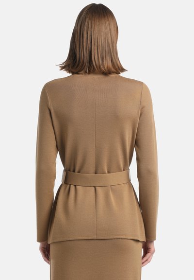 Femme aux cheveux bruns lisses et longueur épaule, portant un cardigan en tricot camel ceinturé et une jupe assortie, vue de dos.