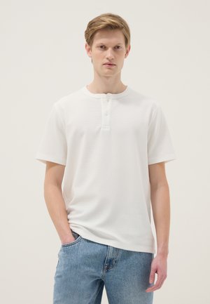 Joven con cabello rubio que lleva una camisa henley blanca de manga corta y jeans azules, de pie con una mano en el bolsillo contra un fondo liso.