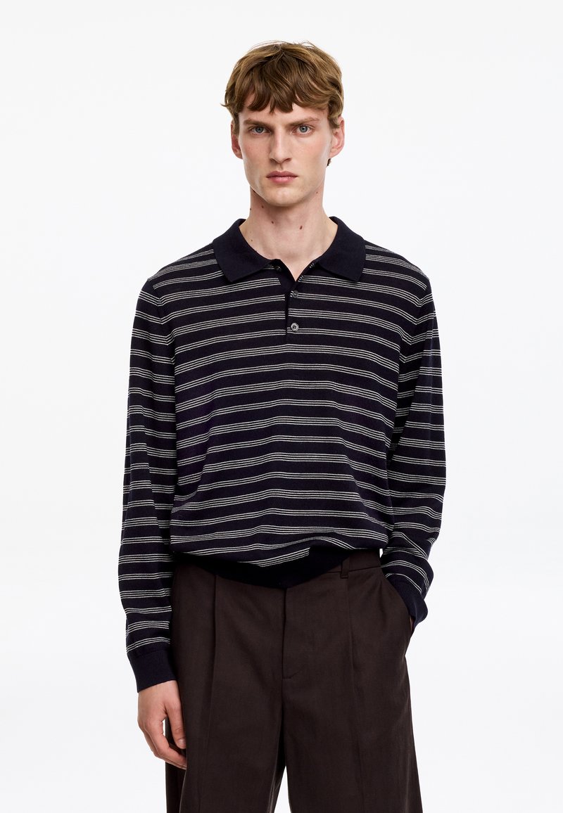 Jeune homme aux cheveux châtain clair courts, portant un polo noir à manches longues avec des rayures blanches horizontales et un pantalon foncé, debout avec une main dans la poche.
