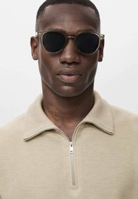 Gafas de sol de montura redonda con lentes oscuros y montura de plástico transparente. El modelo lleva una camiseta de cuello beige con cremallera y tejido texturizado.