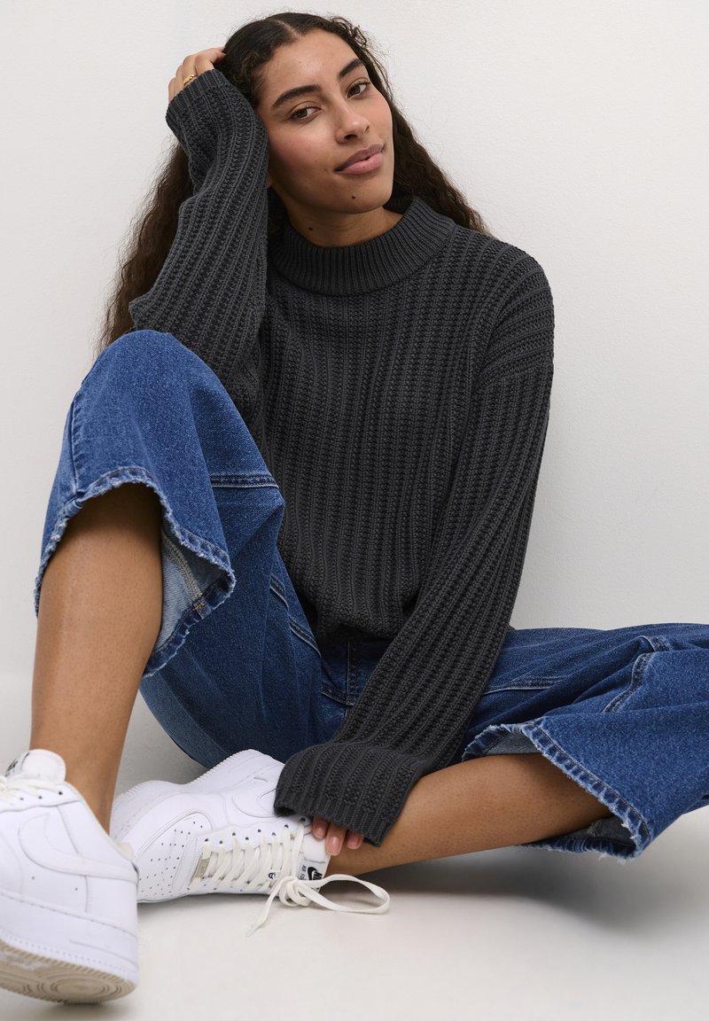 Ciemnoszary sweter z prążkowanego dzianiny z wysokim kołnierzem, zestawiony z szerokimi, jasnoniebieskimi dżinsami i białymi trampkami, siedzący na tle w białym kolorze.