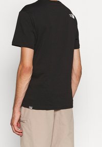 Black cotton t-shirt featuring a crew neck and short sleeves, with a small logo on the upper back. Paired with light beige shorts.