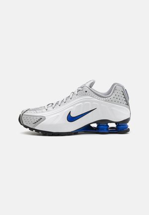 Biały i szary but sportowy z niebieskim logo Nike, czarną podeszwą oraz widoczną niebieską sprężynową amortyzacją w okolicy pięty.