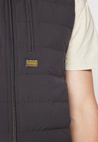 Vadderad svart väst med horisontell sömnad, framzipper och G-Star Originals-logotyp patch i gult. Bärs över en beige skjorta.