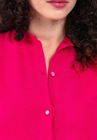 Nahaufnahme der Lippen, des Halses, der lockigen Haare einer Frau und eines leuchtend pinken, mit einem Knopf geschlossenen Pullovers.