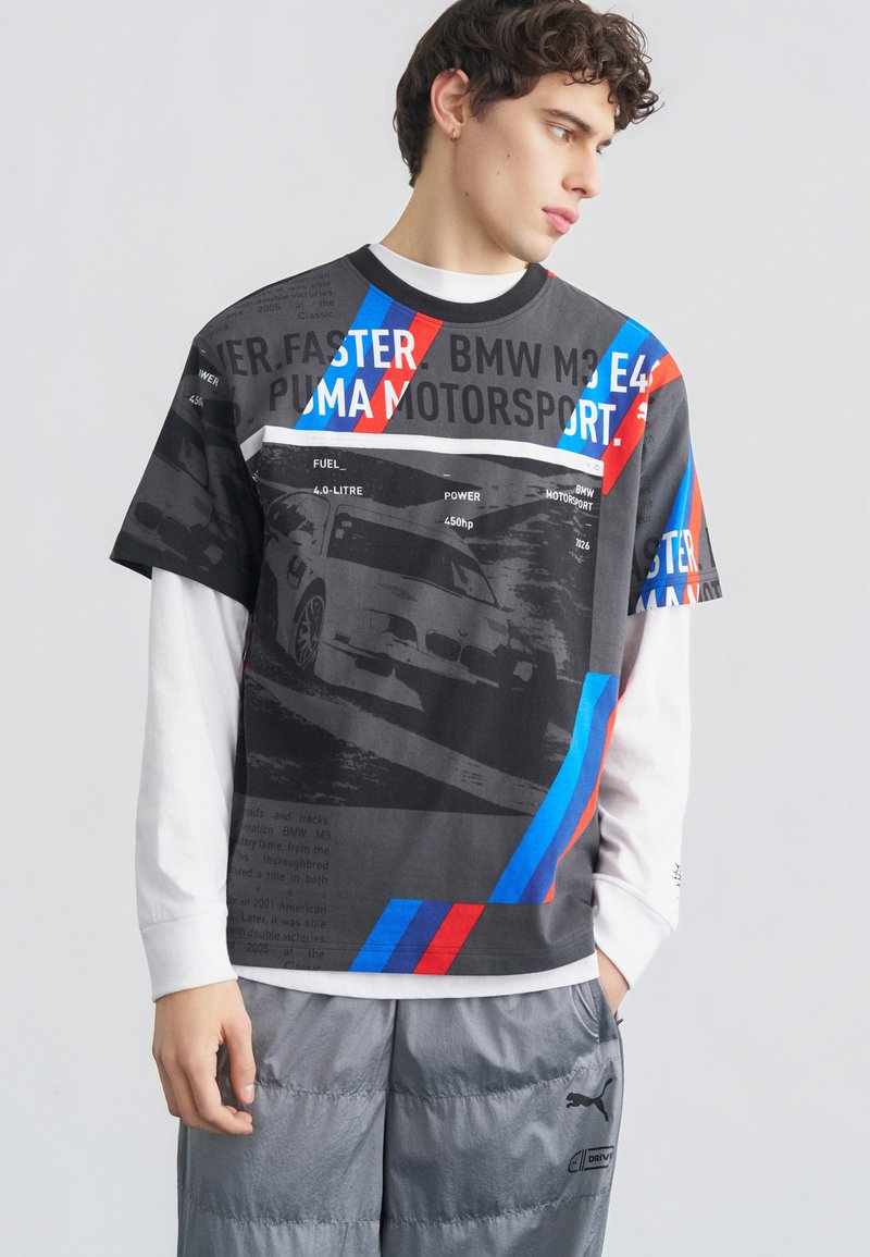 Νέος άνδρας που φοράει μπλουζάκι BMW Motorsport με γραφικά πάνω από λευκή μπλούζα μακρυμάνικη και γκρι παντελόνι Puma, κοιτάζοντας προς τα πλάγια.