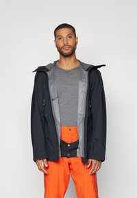 Waterdichte zwarte jas met een grijze binnenkant, voorzien van ritszakken en een capuchon, gedragen over een grijs shirt en oranje broek.