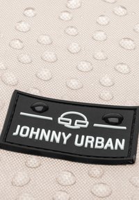 Fekete gumicímke fehér "JOHNNY URBAN" felirattal egy strukturált, vízlepergető bézs háttéren, amelyet tiszta cseppek tarkítanak.