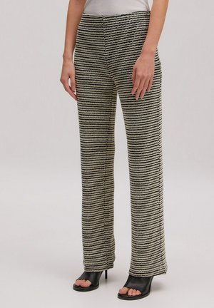 Frau trägt schwarz-weiß gestreifte Strickhose und schwarze offene High-Heel-Sandalen, steht mit locker an den Seiten hängenden Händen.