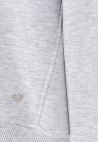 Sweatshirt gris en tissu doux et texturé. Présente un petit logo argenté sur l'ourlet et des poignets côtelés le long du bas et des manches.