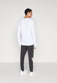 Chemise blanche à manches longues avec une coupe décontractée, associée à un pantalon gris foncé fuselé avec des coutures diagonales et un accent logo blanc. Baskets blanches.