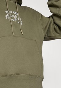 Sweat-shirt olive avec une poche kangourou à l'avant, capuche à cordon de serrage, et un design graphique en blanc sur la poitrine. Tissu doux et texturé.