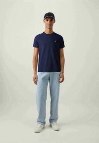 Navy blue cotton t-shirt with a crew neckline and a small yellow logo on the left chest, paired with light blue jeans and white sneakers.