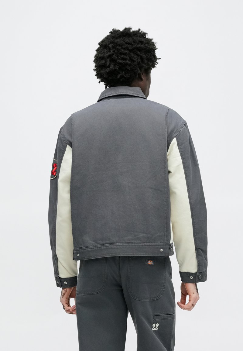 Gray denim jacket with cream-colored sleeves, featuring a button closure, embroidered detail on the arm, and textured fabric.