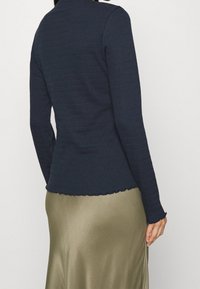 Mörkblå långärmad topp i texturerat stickat material och vågig nederkant, matchad med en slät olivgrön kjol. Bakifrån, händerna placerade vid sidorna.