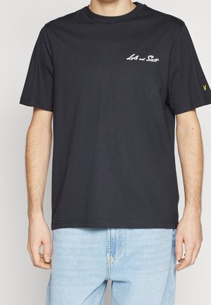 T-shirt noir à manches courtes avec inscription blanche "Lyle and Scott" sur la poitrine et petit logo jaune sur la manche gauche, porté avec un jean bleu clair.