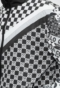 Ύφασμα μπουφάν με ασπρόμαυρο καρό μοτίβο που περιλαμβάνει κυκλικά λογότυπα, λευκό φερμουάρ, αντανακλαστική λωρίδα και διακοσμητικές λεπτομέρειες στα μανίκια.