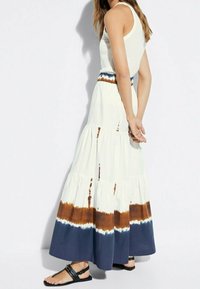 Femme portant une robe blanche sans manches avec des bandes tie-dye bleues et brunes près de l'ourlet, accompagnée de sandales plates noires.