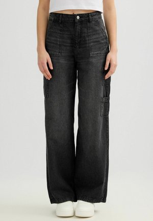 Mujer vestida con pantalones de mezclilla negros de pierna ancha y talle alto, con bolsillos parche delanteros y laterales, combinados con zapatillas blancas y un top blanco corto.